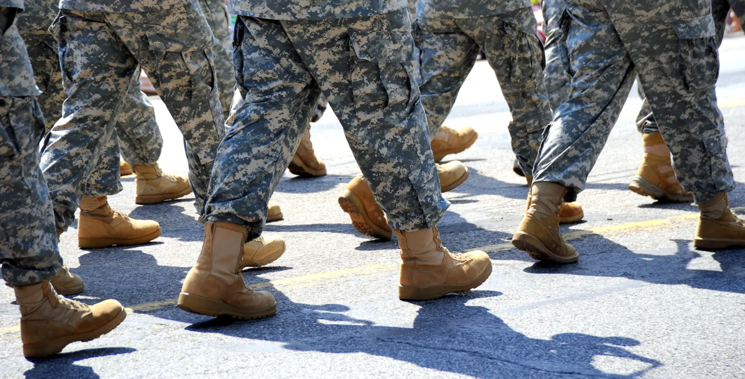 A group of people in military camouflage uniforms and tan boots march in formation on a paved road, reminiscent of the disciplined teamwork valued by any Boston government contract fraud attorney.