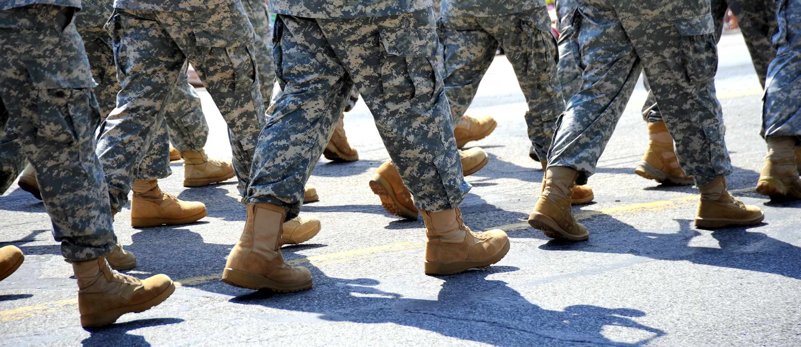 Home A group of people in military camouflage uniforms and tan boots march in formation on a paved road, reminiscent of the disciplined teamwork valued by any Boston government contract fraud attorney.