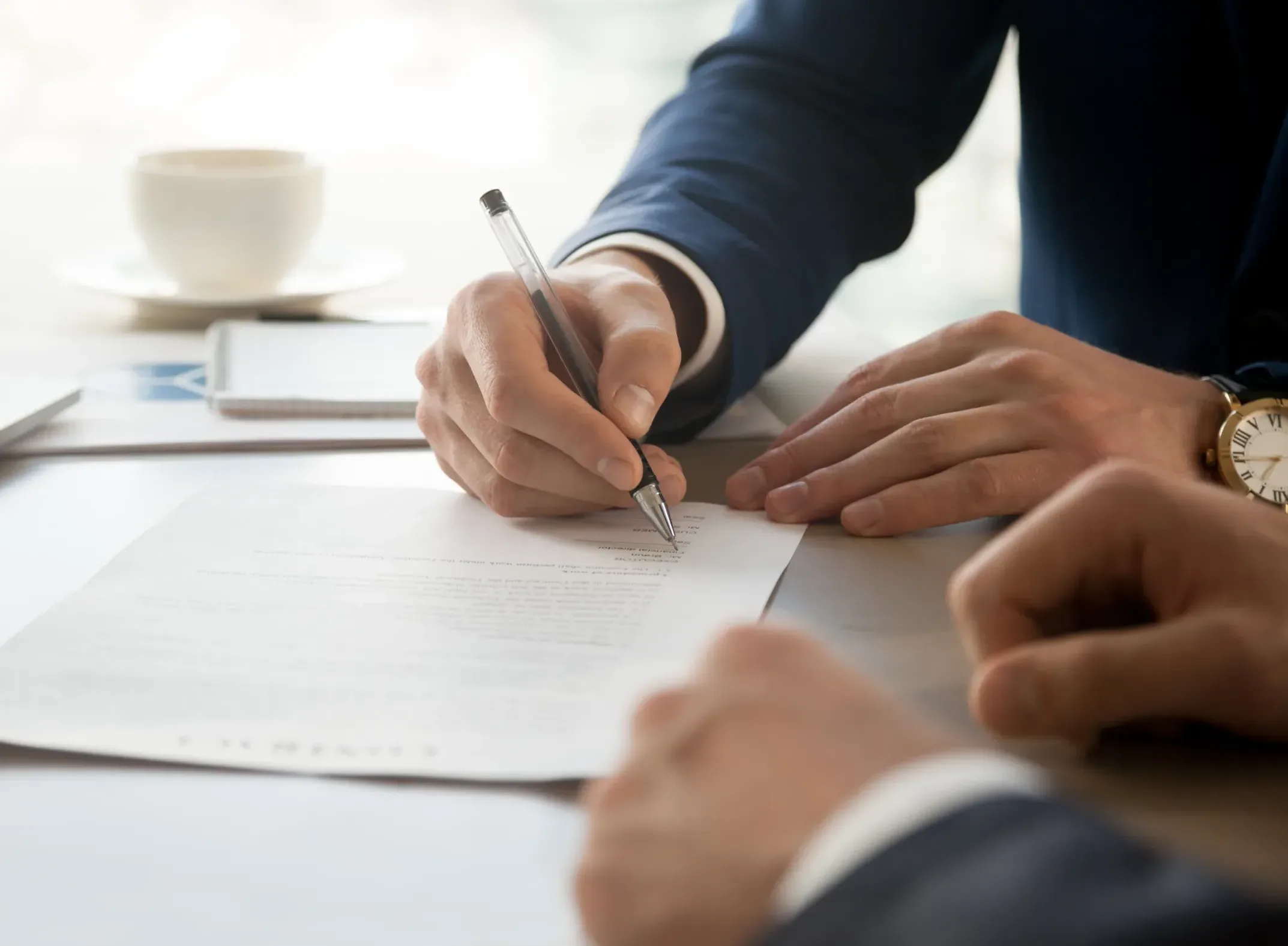 Home Two people in business attire sit at a table; one is signing a document with a pen while the other observes, reflecting the diligence often seen among Silicon Valley CFTC whistleblower attorneys. A cup of coffee and a smartphone are on the table in the background.