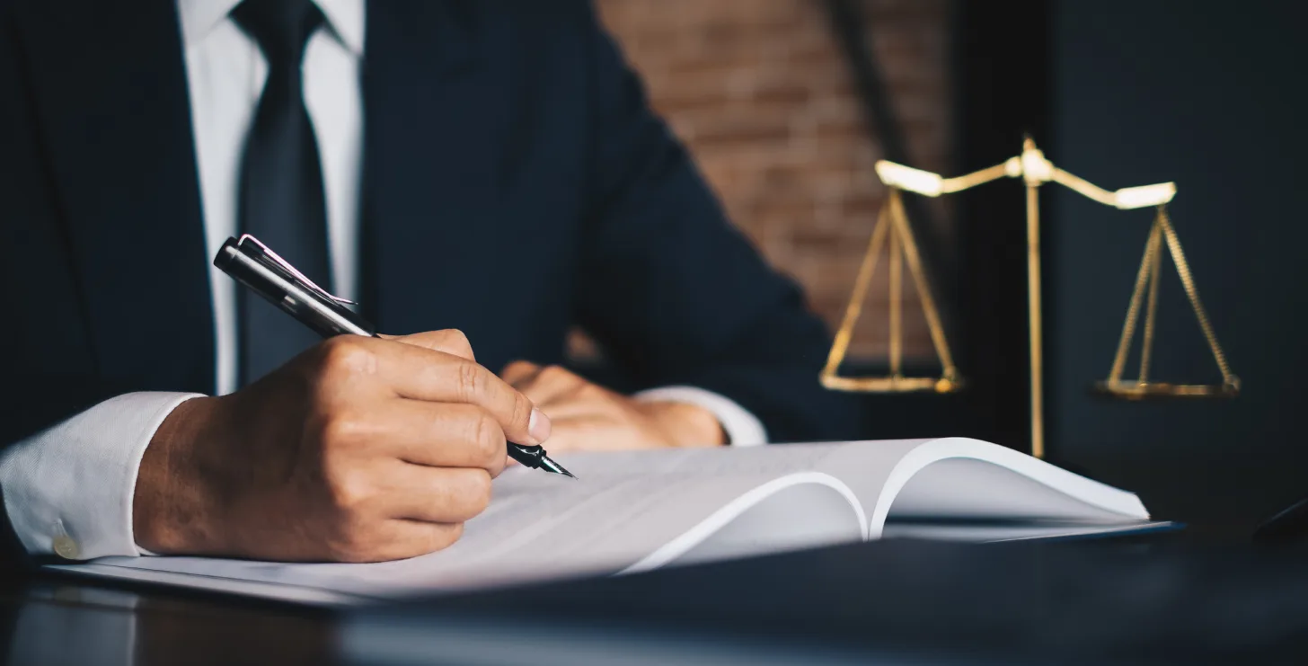 A person in a suit writes in a large open book at a desk, with golden scales of justice in the background—perhaps preparing an important case as a New York Medicare whistleblower.
