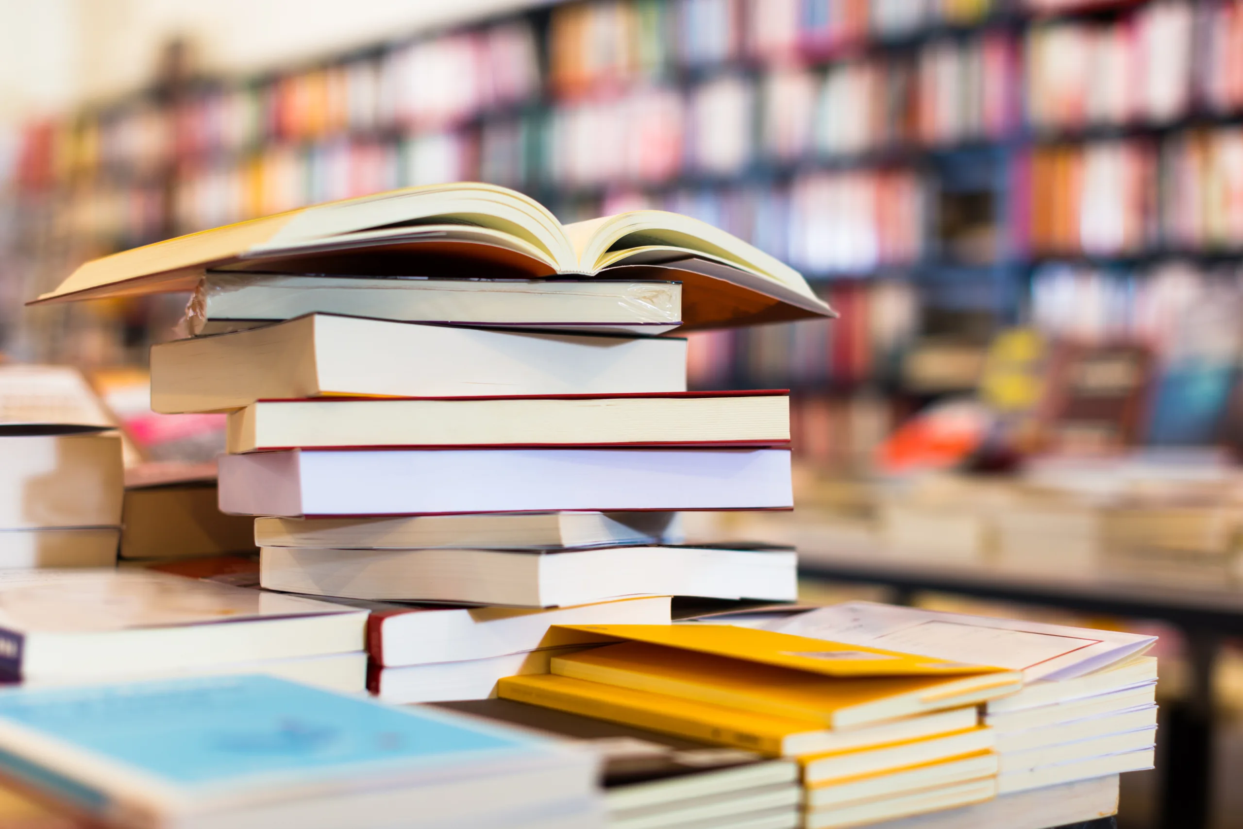 Home A stack of books, with one open on top, sits on a table in a bookstore or library—perhaps awaiting the next reader interested in topics like Boston government contract fraud attorney expertise. Rows of blurred bookshelves are visible in the background.