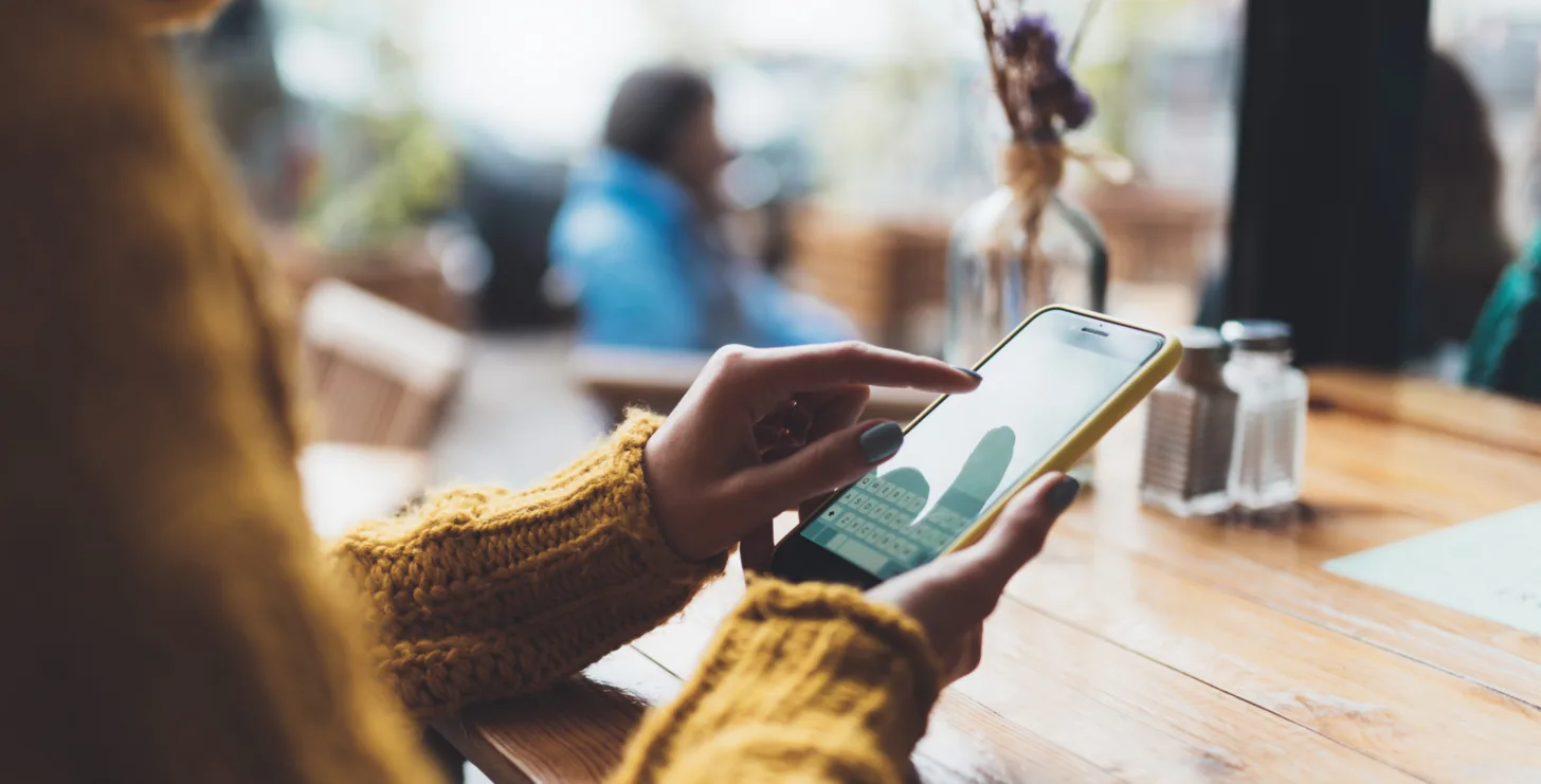 A person in a yellow sweater sits at a wooden table in a café, using a smartphone while blurred people and a vase create a cozy backdrop—perhaps researching San Jose IRS whistleblower lawyers or seeking advice from Silicon Valley CFTC whistleblower attorneys.