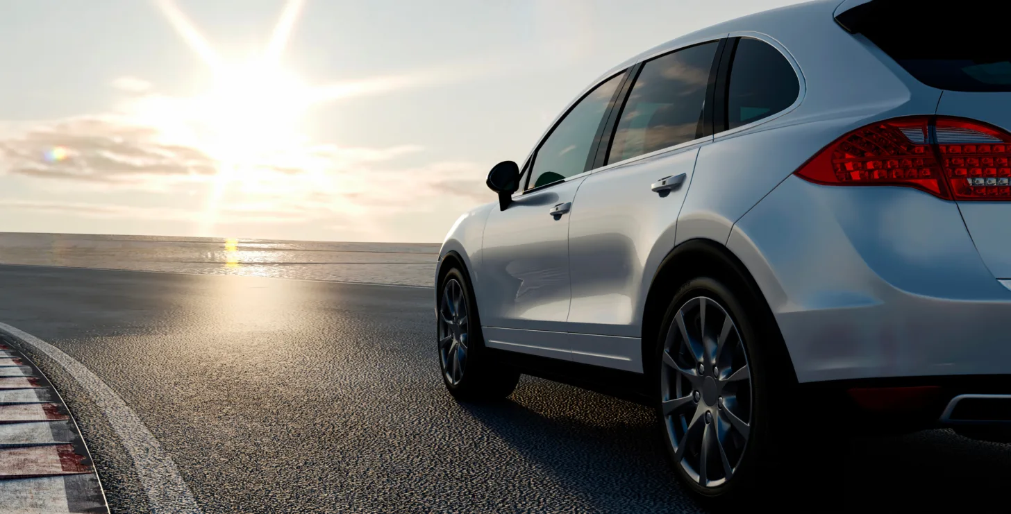 A white SUV is parked on a coastal road at sunset. The sun is low in the sky, reflecting off the water and pavement, much like how critical insights from a New York Medicare whistleblower can shed light on hidden details beneath the surface.