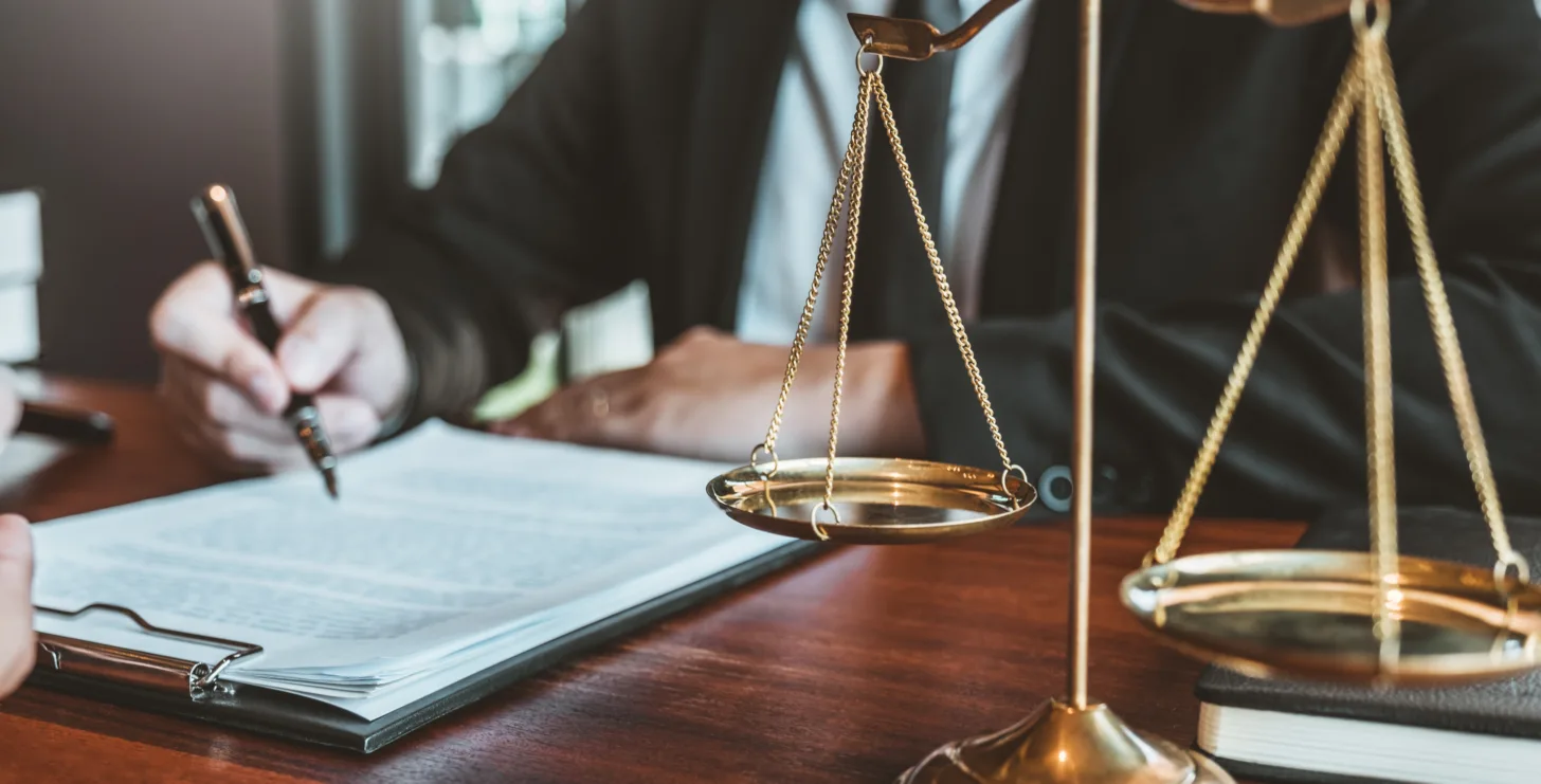 A person in business attire writes on documents at a desk with a brass balance scale and legal books in the foreground, conveying the professionalism and diligence of a New York City tax whistleblower lawyer.