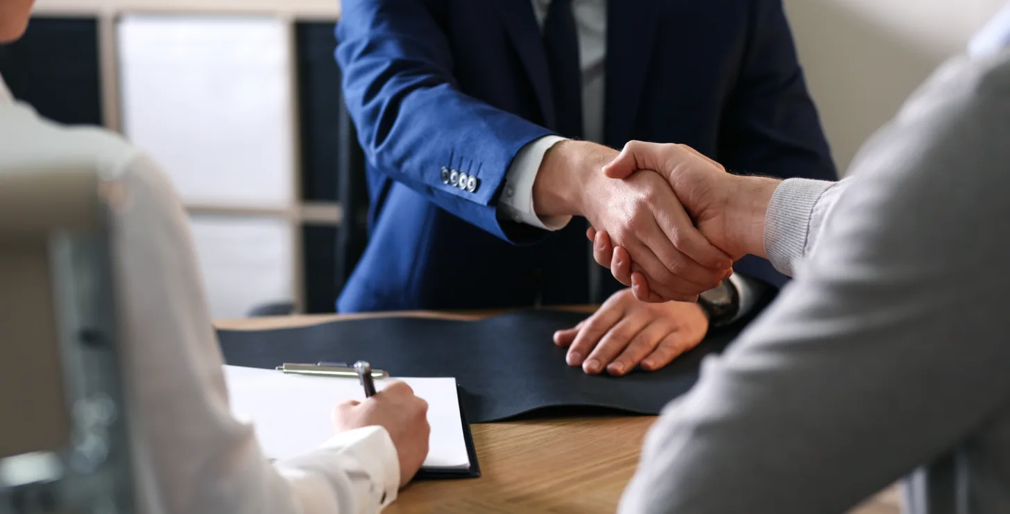 Two people shake hands across a desk while a third person takes notes, suggesting a business meeting or interview in an office setting—an atmosphere where guidance from professionals like a Boston government contract fraud attorney can be essential.