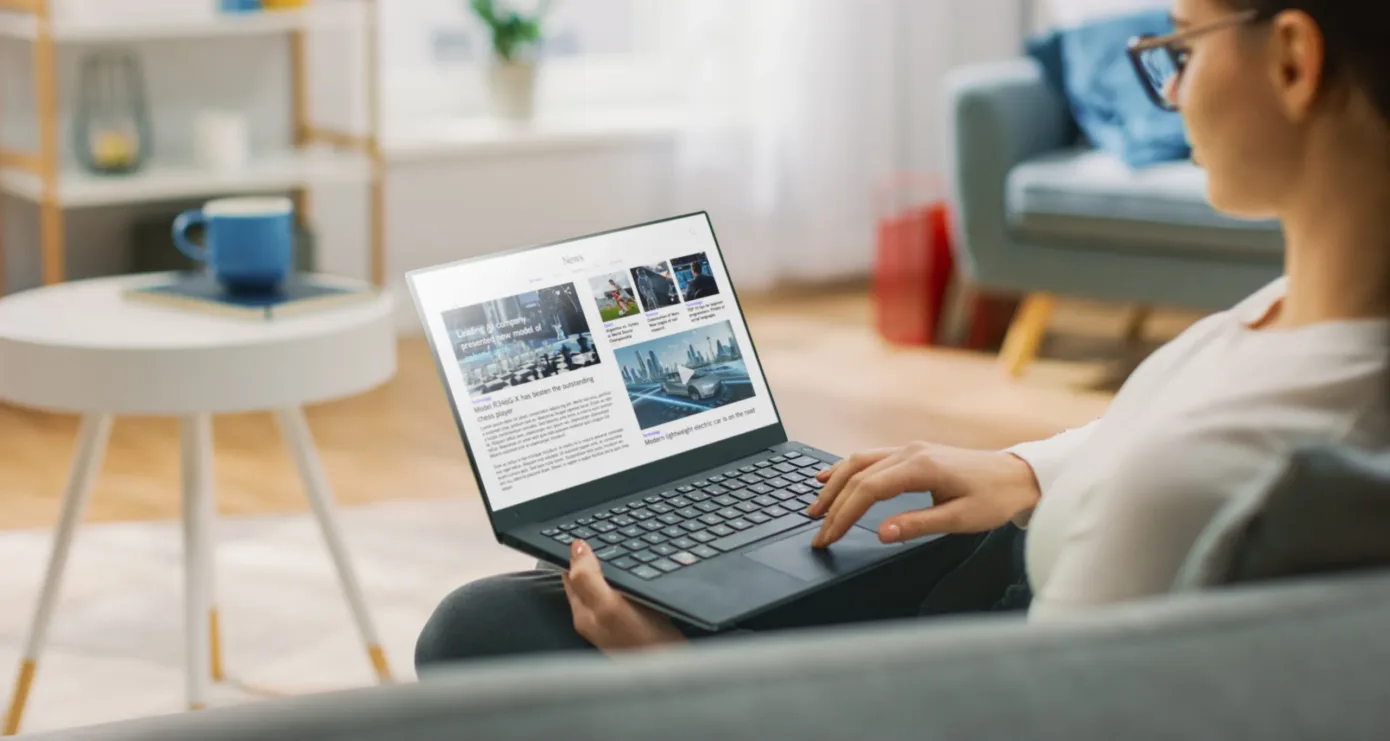 Person sitting on a couch using a laptop, viewing a news website with articles and images displayed on the screen, including insights from New York City tax whistleblower lawyer interviews.