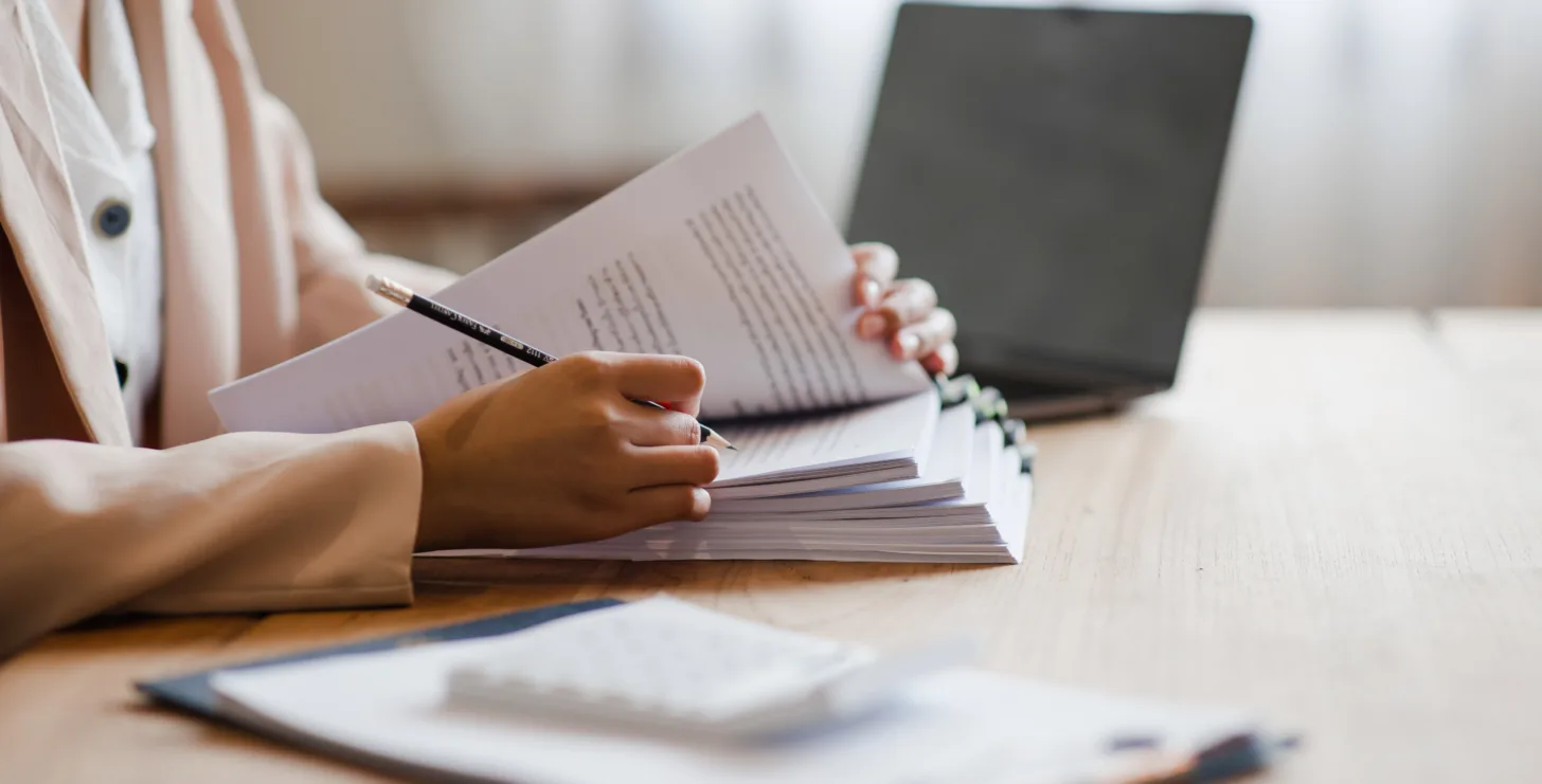 Person sits at a desk, flipping through and marking papers with a pen—perhaps reviewing evidence as a New York City tax whistleblower lawyer would. A laptop, clipboard, and calculator are also on the wooden table.