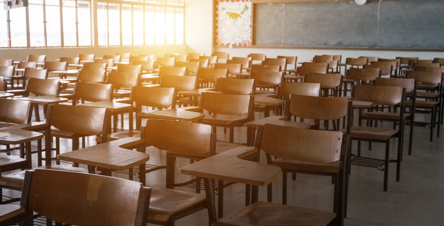 Empty classroom with rows of wooden desks and chairs, sunlight streaming through windows, chalkboard in the background—a quiet space reminiscent of where many Boston government contract fraud attorneys once began their journeys.