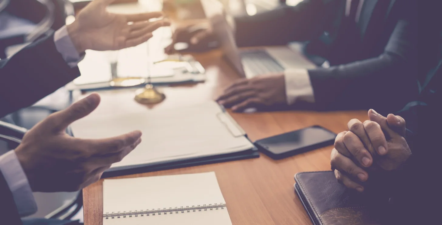 Three people sit at a table with notebooks, a laptop, and documents, engaging in a discussion—only their hands and arms are visible—perhaps consulting with a New York City tax whistleblower lawyer about the details of their case.