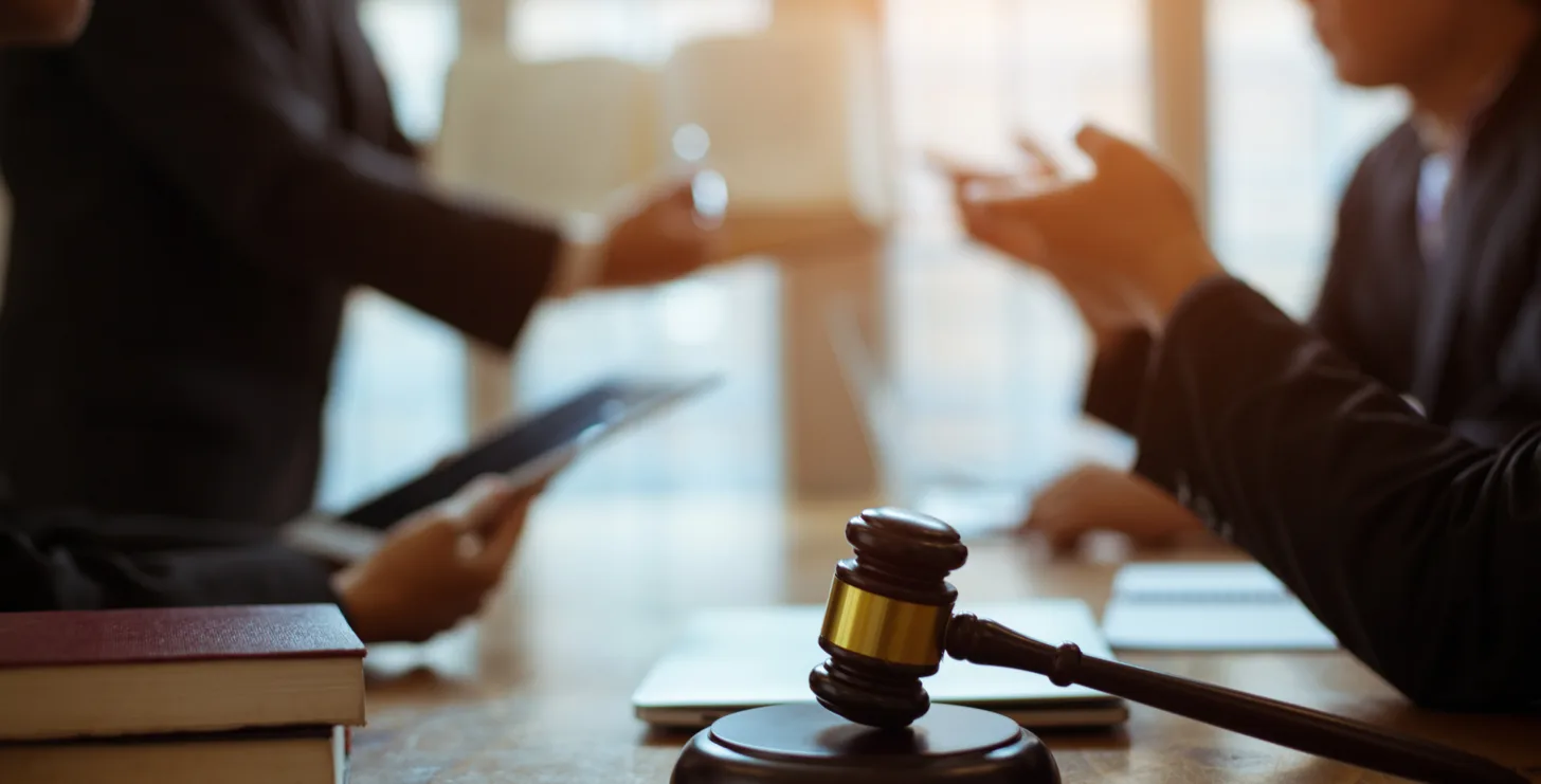 A judge's gavel sits on a table with books and documents, while three people in suits—perhaps San Jose IRS whistleblower lawyers—discuss legal matters in the background.