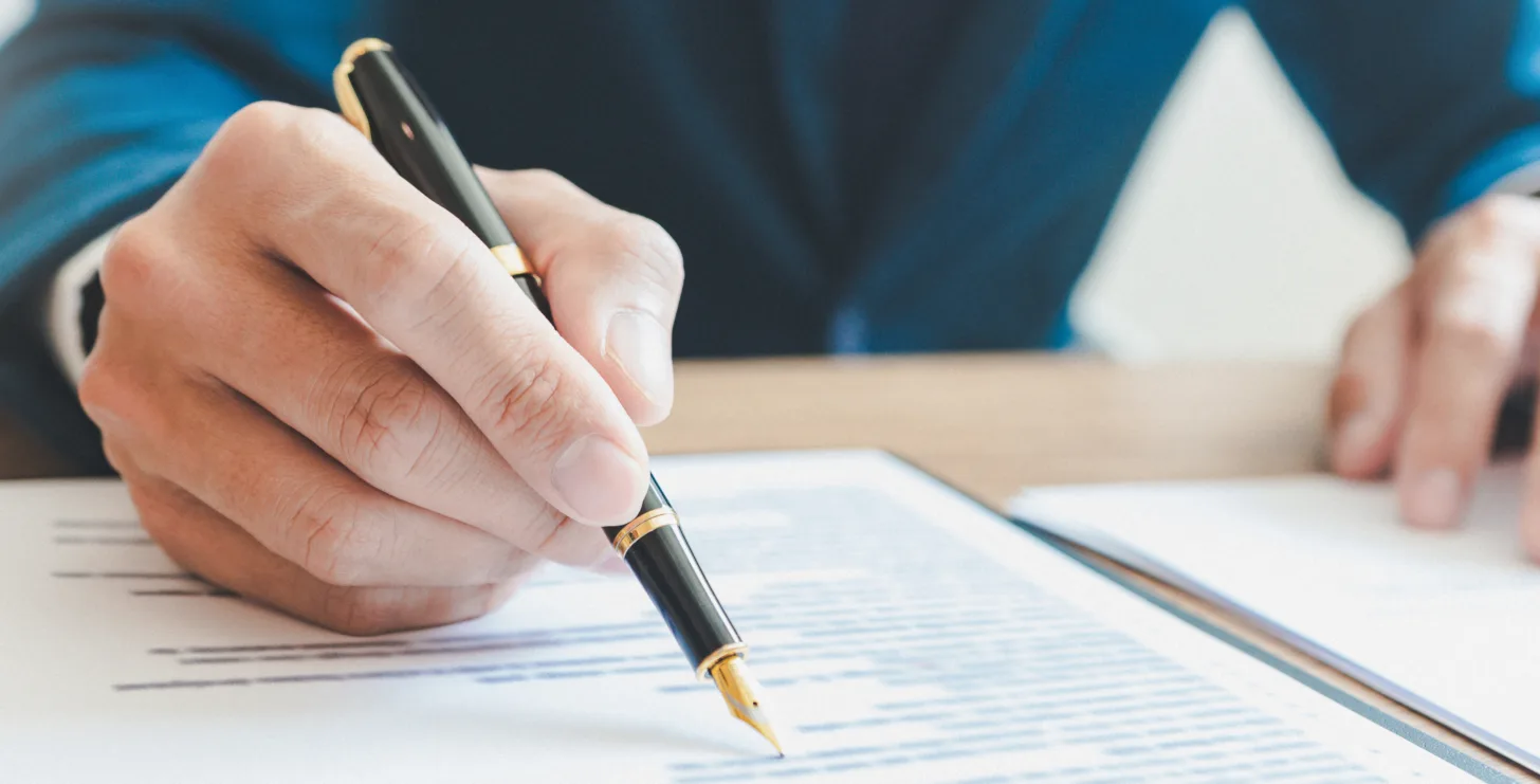 A New York City tax whistleblower lawyer in a suit signs a document with a black and gold pen at a desk.