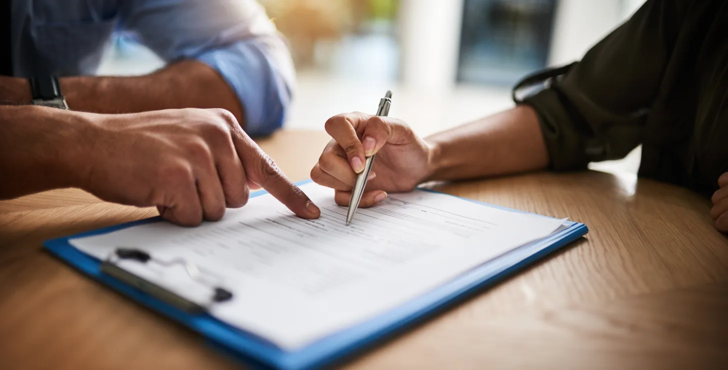 Two people sit at a table; one person points at a document on a clipboard while the other prepares to sign it with a pen, illustrating the careful guidance San Jose IRS whistleblower lawyers provide during sensitive legal matters.