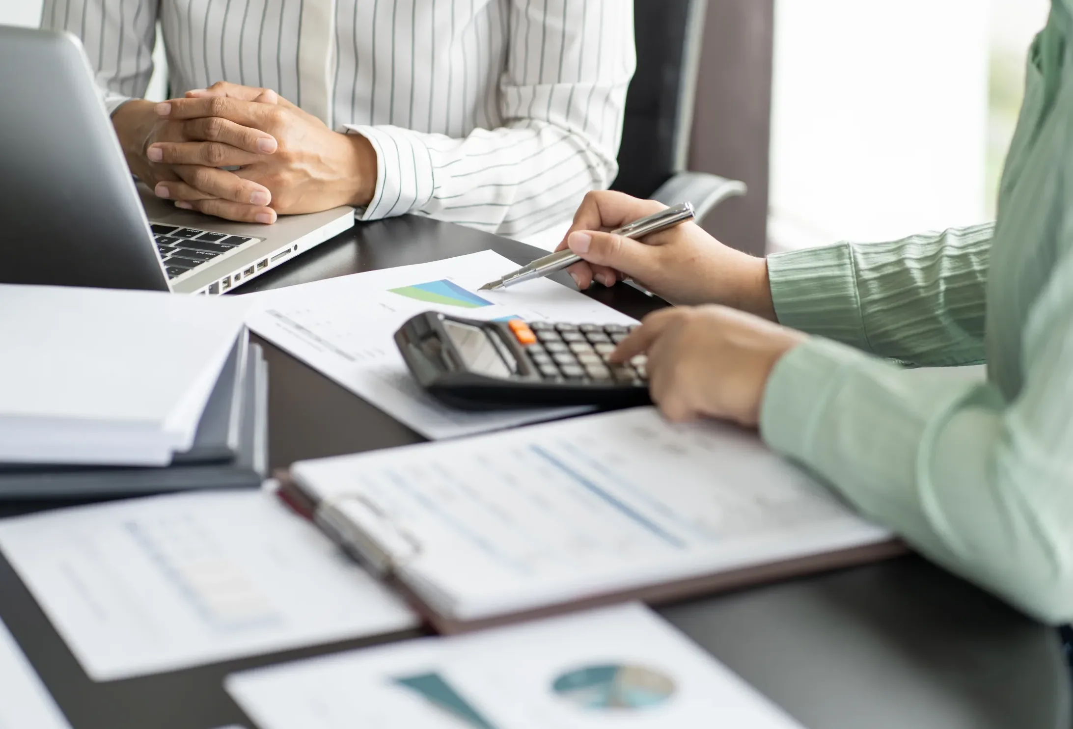 Home Two people review financial documents at a desk, reminiscent of the careful analysis performed by a San Francisco healthcare fraud lawyer. One person uses a calculator, while the other sits with folded hands near a laptop. Charts and reports are spread out on the table.