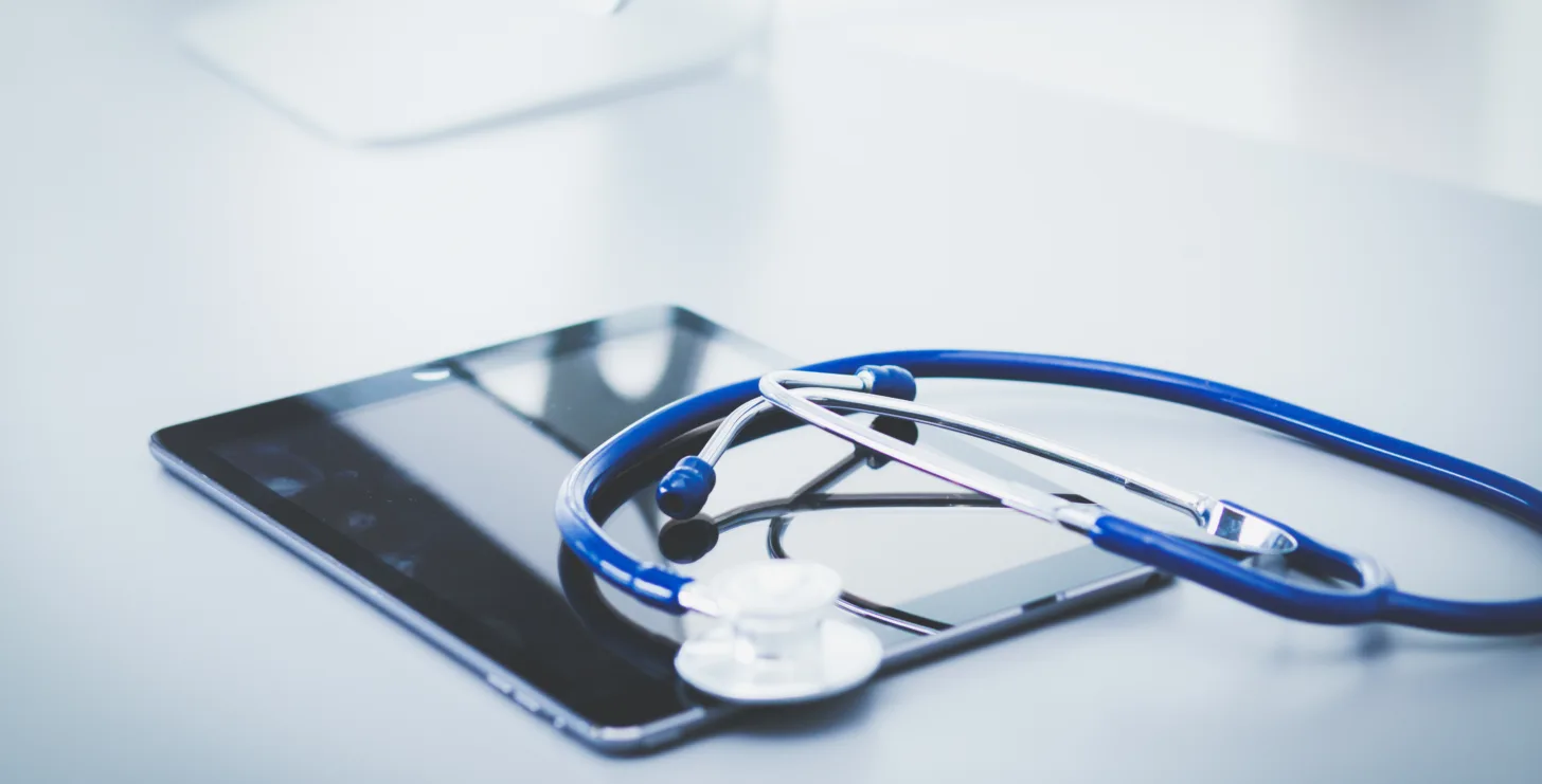 A blue stethoscope rests on top of a tablet device on a white desk, suggesting a connection between medicine and technology—much like how a New York Medicare whistleblower bridges the gap between healthcare and legal oversight.