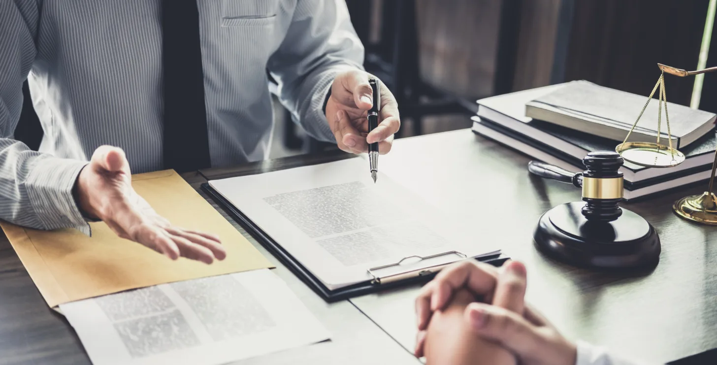 Two people sit at a desk discussing documents. Legal books, a gavel, and scales of justice are on the table, suggesting a legal or business meeting—perhaps between a New York City tax whistleblower lawyer and their client navigating critical compliance matters.
