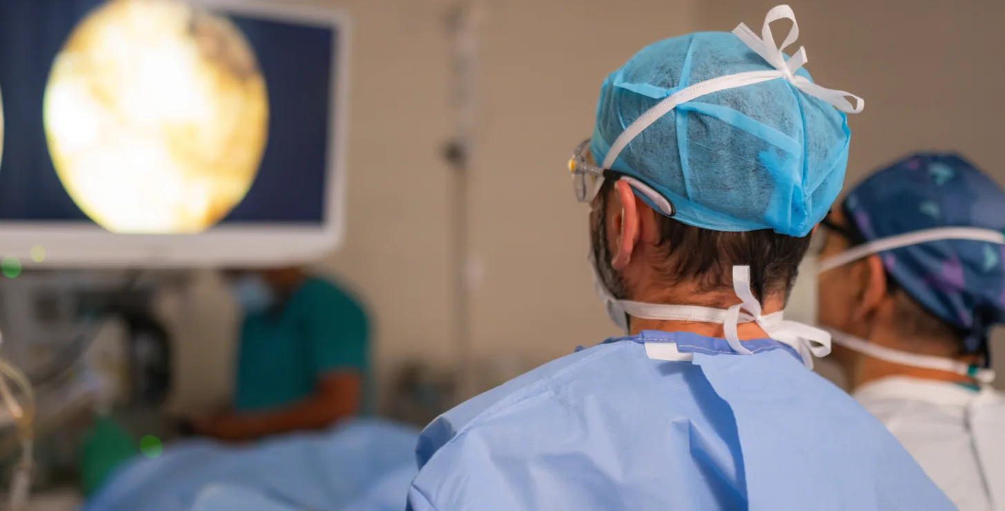 Two surgeons in blue gowns and caps perform a medical procedure while viewing a monitor displaying an internal image in an operating room, reminiscent of cases investigated by a New York Medicare whistleblower dedicated to exposing healthcare fraud.