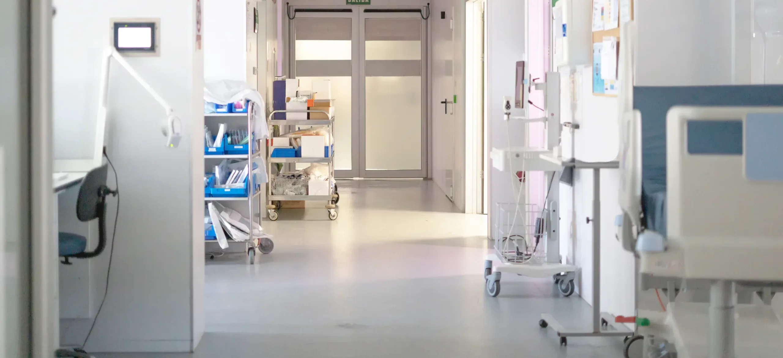 Home A hospital corridor with medical equipment, carts, and beds along the walls sets a professional scene reminiscent of the environments investigated by a San Francisco healthcare fraud lawyer. Double doors are visible at the end of the brightly lit hallway, emphasizing both security and accessibility within healthcare facilities.