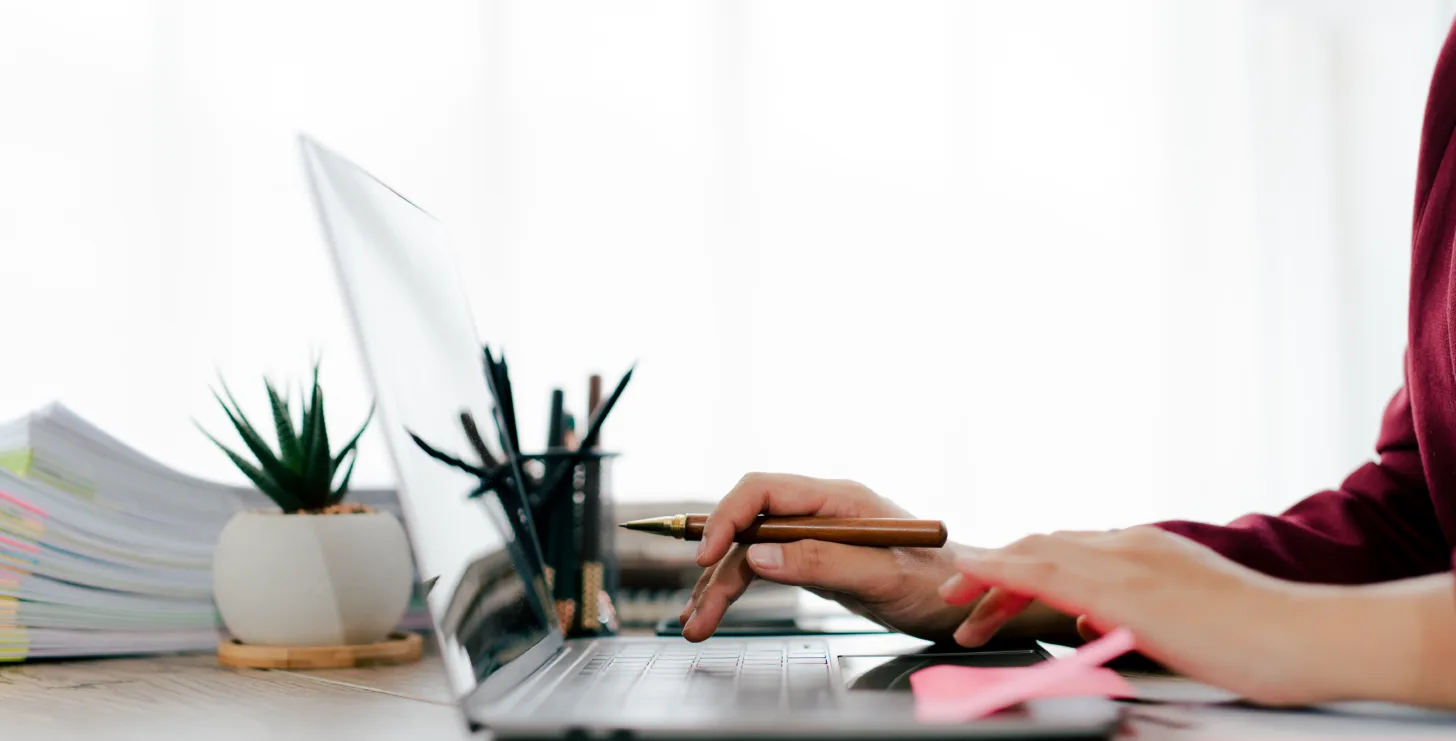 Person using a laptop at a desk with a potted plant, pen holder, stacked papers, and sticky notes in a bright workspace—a scene reminiscent of the organized environments where San Jose IRS whistleblower lawyers or Silicon Valley CFTC whistleblower attorneys might prepare important case documents.