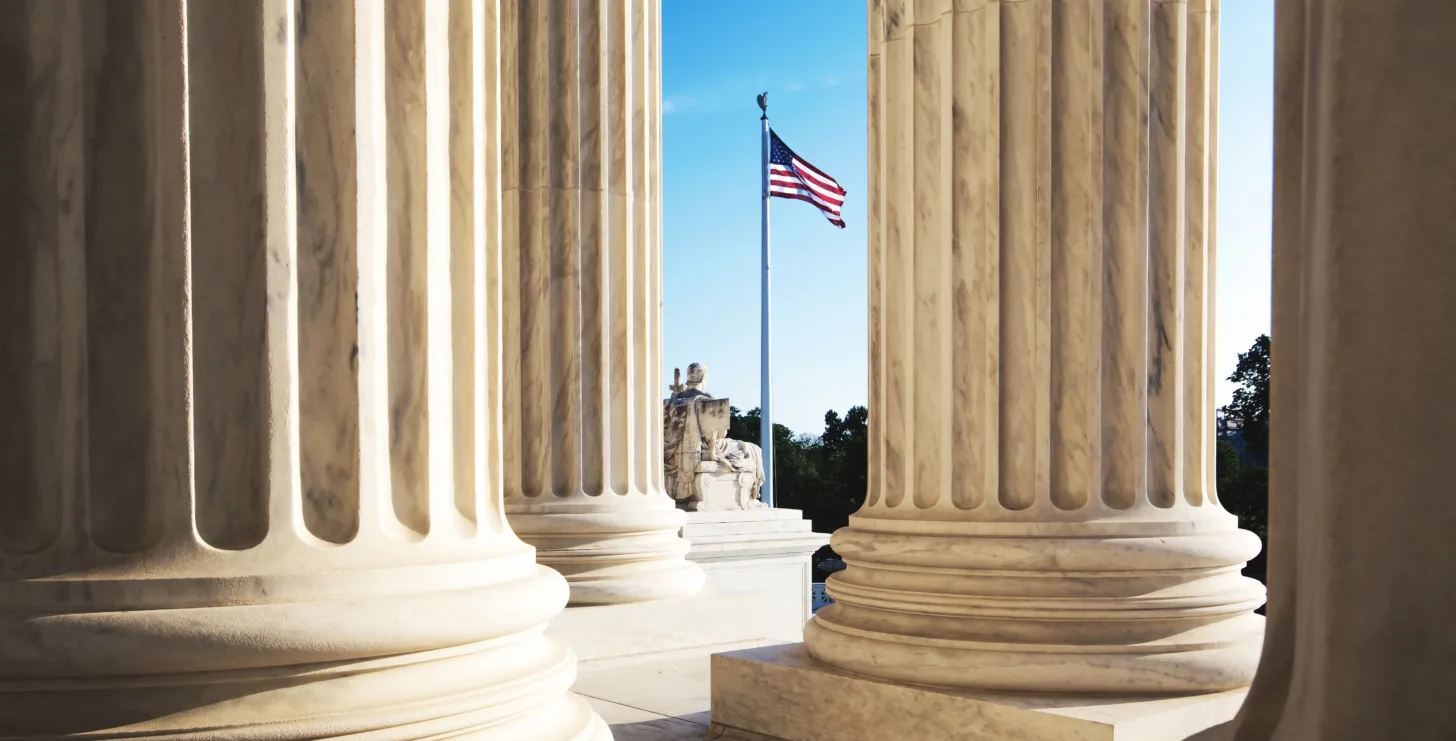 Large marble columns frame a view of an American flag flying outside on a sunny day, with a statue visible in the background—a scene reminiscent of the steadfast integrity embodied by New York City tax whistleblower lawyers.