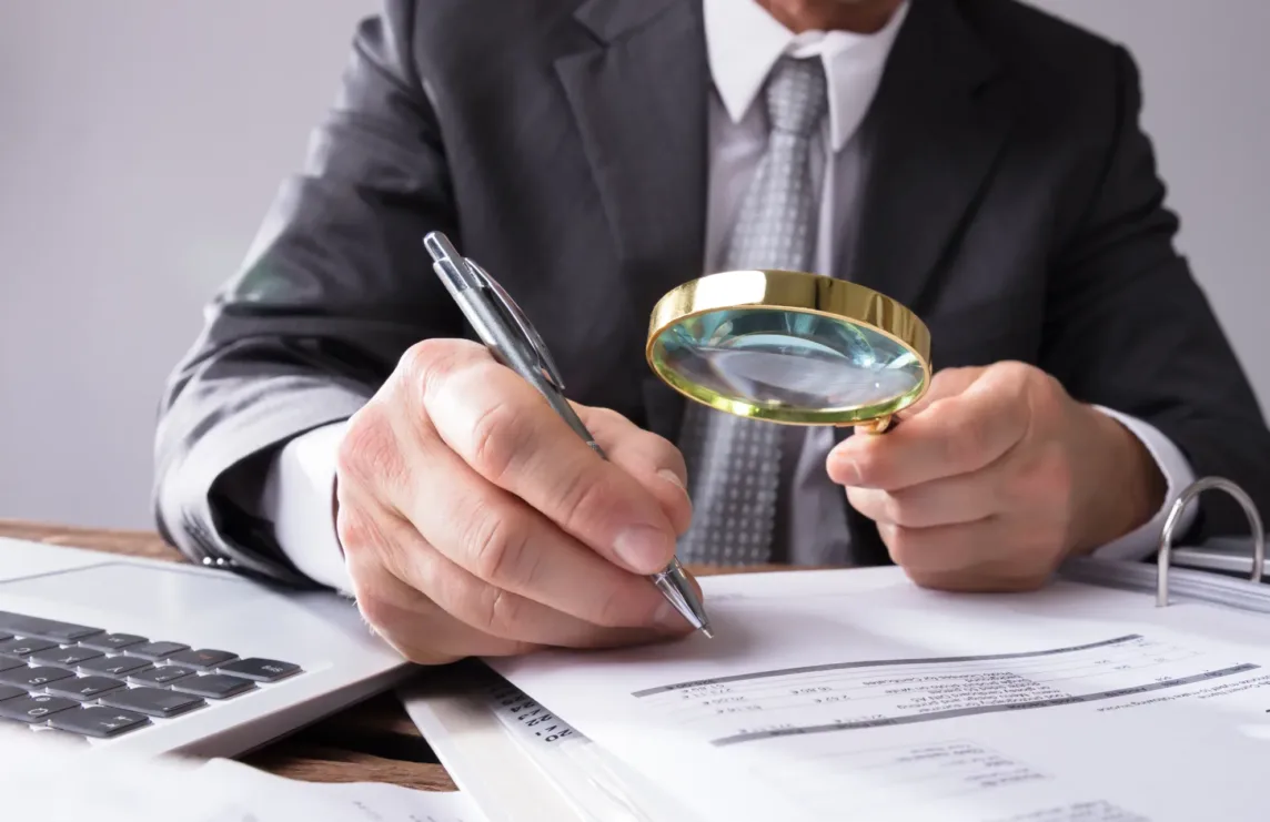 A person in a suit examines financial documents with a magnifying glass while holding a pen, next to a calculator and laptop on a desk—a scene reminiscent of the meticulous review undertaken by San Jose IRS whistleblower lawyers.