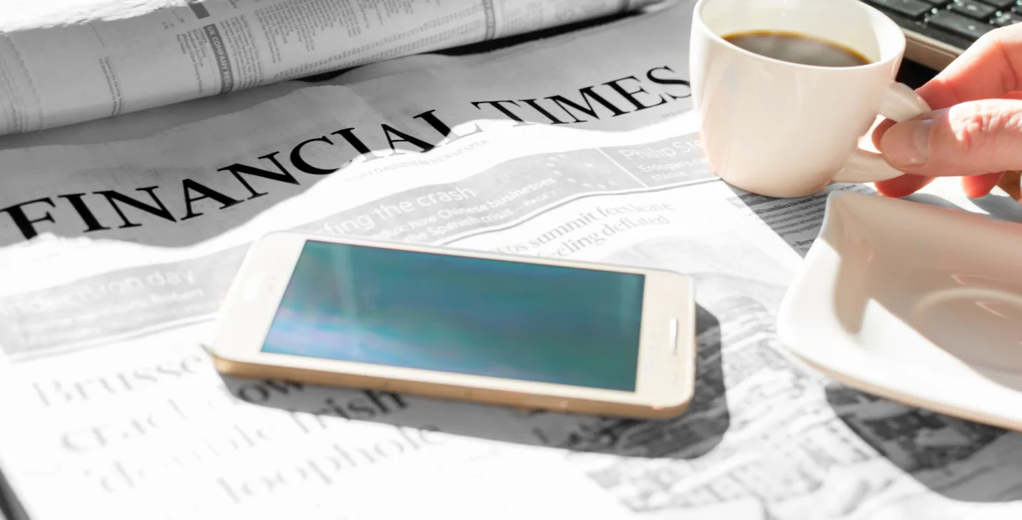 A person holds a cup of coffee next to a smartphone and a Financial Times newspaper on a table—perhaps starting their day by reading the headlines, or preparing to consult with their New York City tax whistleblower lawyer about recent financial developments.
