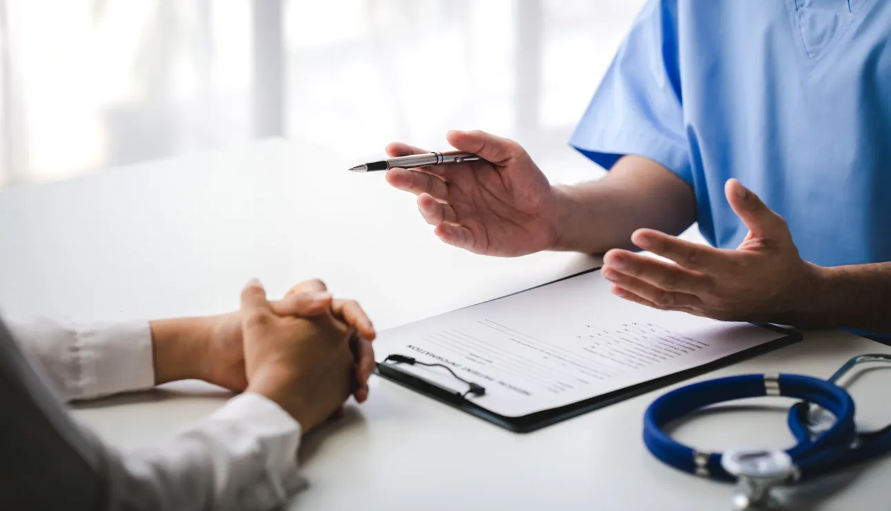 Doctor discussing medical information with a patient at a desk, with a clipboard, pen, and stethoscope visible—an important setting where concerns about healthcare compliance may arise. In cities like San Francisco, a healthcare fraud lawyer or even a New York Medicare whistleblower could play pivotal roles in addressing potential issues of fraud or misconduct within clinical environments.