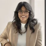 A woman with wavy dark hair and glasses, wearing a tan blazer over a white top, sits and smiles at the camera in a well-lit room—her confident demeanor reminiscent of a dedicated San Francisco healthcare fraud lawyer.