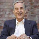 A man in a dark blazer and white shirt sits and smiles in front of a brick wall background, reminiscent of a confident New York Medicare whistleblower ready to take on new challenges.