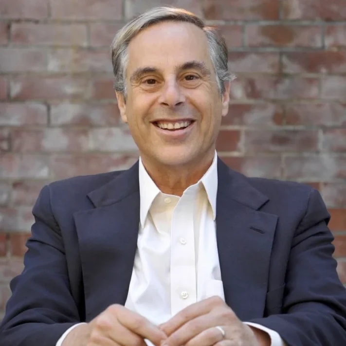 A man in a dark blazer and white shirt sits and smiles in front of a brick wall background, reminiscent of a confident New York Medicare whistleblower ready to take on new challenges.