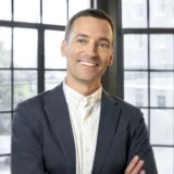 Man in a gray blazer and white shirt stands in front of large windows, smiling with arms crossed, in a bright modern office setting—reflecting the confidence and professionalism of a leading San Francisco healthcare fraud lawyer.