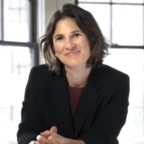 A woman with short brown hair in a black blazer sits at a table, smiling, with large windows and natural light in the background—much like you might find in the offices of top Silicon Valley CFTC whistleblower attorneys or San Jose IRS whistleblower lawyers.