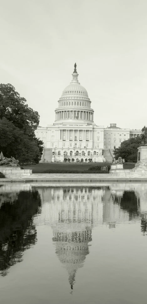 The United States Capitol building is reflected in a calm pool of water, surrounded by trees, in a black and white photograph—a striking image that resonates with those who seek justice, such as a Boston government contract fraud attorney.