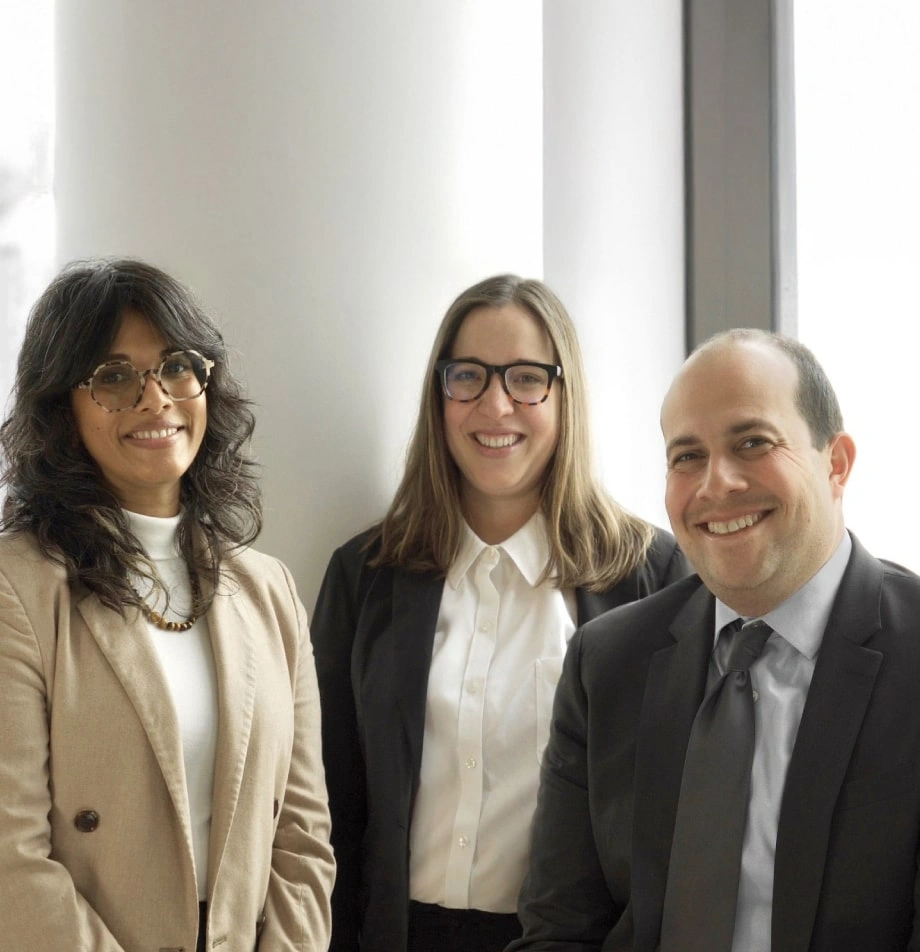 Three people in business attire stand indoors, smiling at the camera. Two women wear glasses and blazers; one man, who could easily be mistaken for a Boston government contract fraud attorney, wears a suit and tie. Light enters through a nearby window.