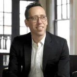 Man in a black suit and white shirt sits indoors on a chair, smiling, with large windows and natural light in the background—exuding the professionalism you’d expect from a seasoned San Francisco healthcare fraud lawyer.