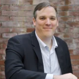 A man in a dark blazer and checkered shirt sits in front of a brick wall, smiling slightly at the camera—a look that reflects the confidence and composure often found in a seasoned New York City tax whistleblower lawyer.