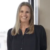 A woman with long blonde hair, wearing a black blouse, stands indoors by a window and smiles at the camera—reminiscent of the dedicated professionalism displayed by Silicon Valley CFTC whistleblower attorneys who confidently advocate for their clients.