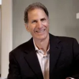 A man wearing a dark blazer and light shirt smiles while seated indoors against a neutral background, evoking the professional confidence of a New York City tax whistleblower lawyer.