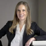 A woman with long blonde hair wearing a black blazer and white blouse smiles at the camera while leaning on a white surface, reflecting the professionalism you can expect when consulting a New York City tax whistleblower lawyer.