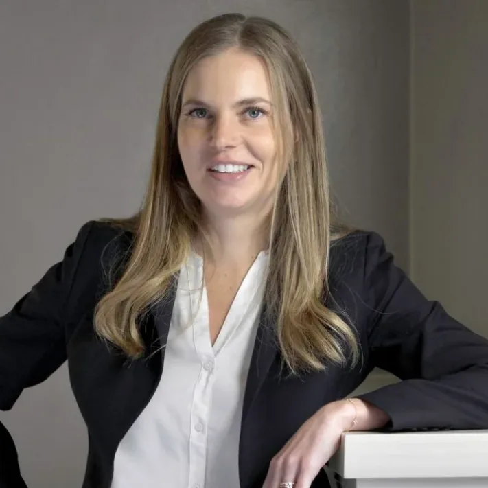 A woman with long blonde hair wearing a black blazer and white blouse smiles at the camera while leaning on a white surface, reflecting the professionalism you can expect when consulting a New York City tax whistleblower lawyer.