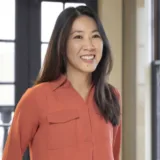 A woman with long dark hair, wearing a rust-colored button-up shirt, stands indoors in front of large windows, smiling—her confident demeanor reminiscent of a dedicated New York City tax whistleblower lawyer.
