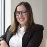 Woman with straight brown hair and glasses, wearing a white blouse and black blazer, smiling while seated indoors near a window—an accomplished New York City tax whistleblower lawyer known for her expertise and dedication.