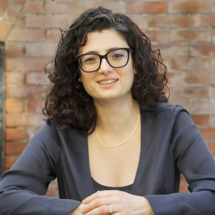 A woman with curly dark hair, glasses, and a dark blouse sits at a table in front of a brick wall, looking at the camera and smiling slightly—her poised demeanor reflecting the confidence seen in leading Silicon Valley CFTC whistleblower attorneys.