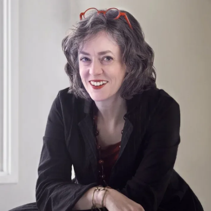 A woman with wavy gray hair, wearing red glasses on her head, a black jacket, and a red top, smiles while leaning slightly forward in a softly lit room—the confident demeanor of someone who could offer trusted advice as a New York City tax whistleblower lawyer.