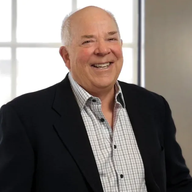 Smiling older man with short white hair wearing a dark blazer and checked shirt, standing in front of a bright window—he exudes the confidence of a seasoned New York Medicare whistleblower.