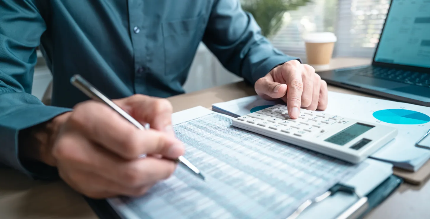 Person using a calculator and writing on a financial spreadsheet at a desk, with a laptop and coffee cup in the background, perhaps reviewing documents for a San Francisco healthcare fraud lawyer or consulting with a Boston government contract fraud attorney.