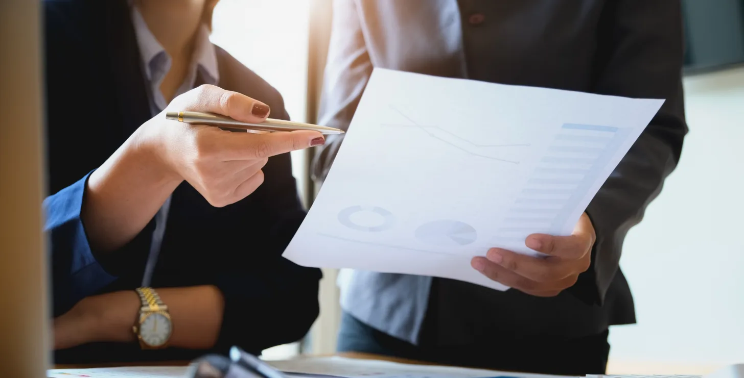 Two people in business attire review a printed sheet with graphs and charts in an office setting, with one person pointing at the paper with a pen—an environment reminiscent of consultations held by experienced San Francisco healthcare fraud lawyers.