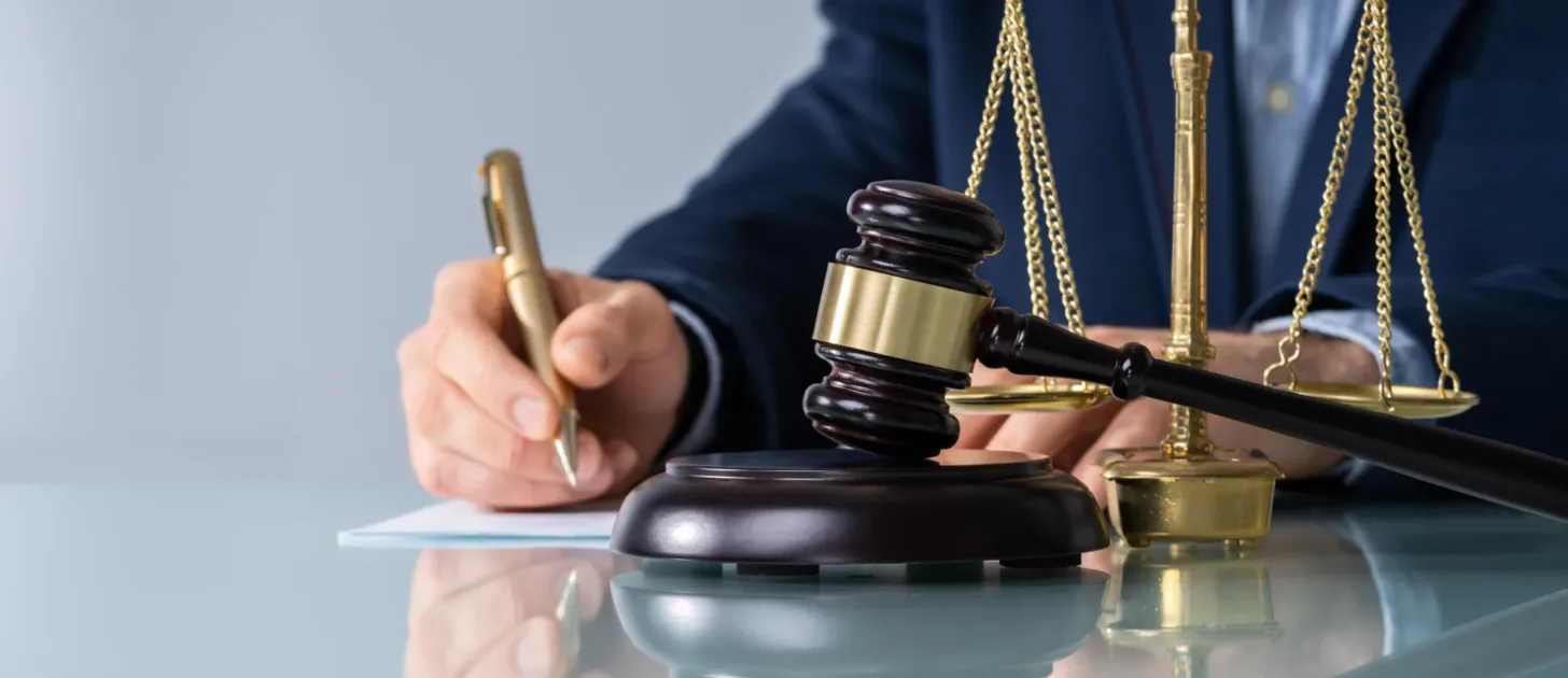 A person writes on paper beside a wooden gavel and scales of justice on a glass desk, suggesting a legal or courtroom setting—perhaps consulting with a New York City tax whistleblower lawyer or discussing important documents related to IRS cases.