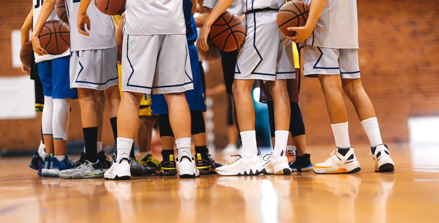 Group of basketball players in uniforms standing in a gym huddle, each holding a basketball, with focus on their legs and shoes—much like a team prepared to tackle challenges together, just as a Boston government contract fraud attorney stands ready to support clients facing legal obstacles.