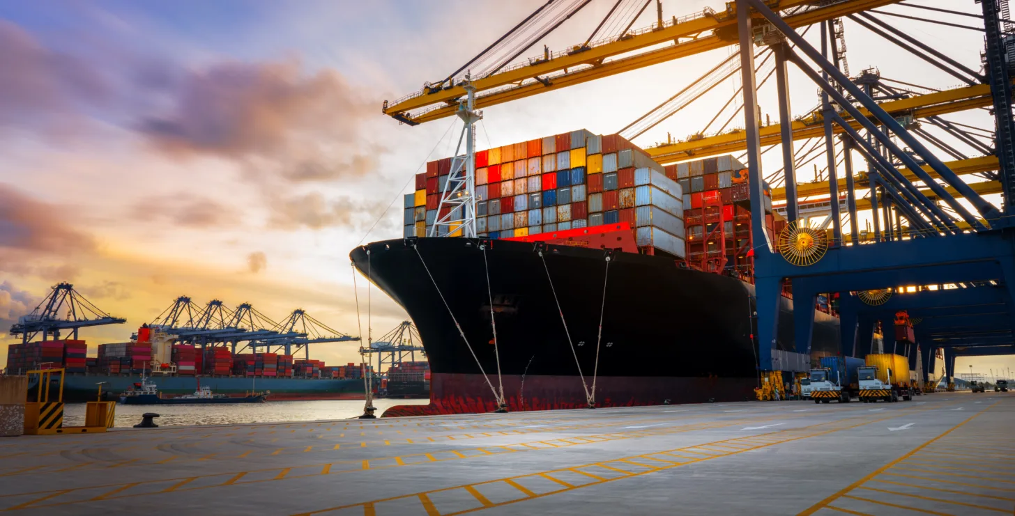 A large cargo ship loaded with colorful shipping containers is docked at a port, with cranes and industrial equipment in the background at sunset—much like the bustling docks where a Boston government contract fraud attorney might investigate complex shipping compliance cases.