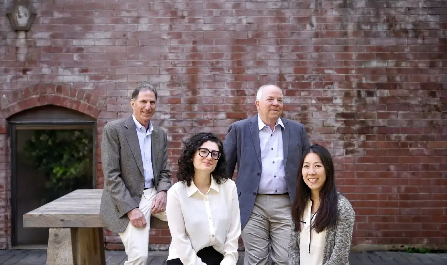 Four adults, two men and two women, pose together in business casual attire in front of a red brick wall outdoors, reminiscent of a dedicated New York Medicare whistleblower team.